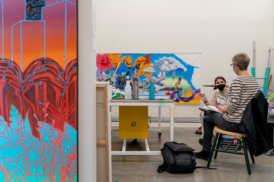 Candid photo of two people in a visual art studio space chatting. A vibrantly-colored painting hangs outside the studio, a desk and chair can be seen inside, as well as more in-progress work pinned to the interior walls.