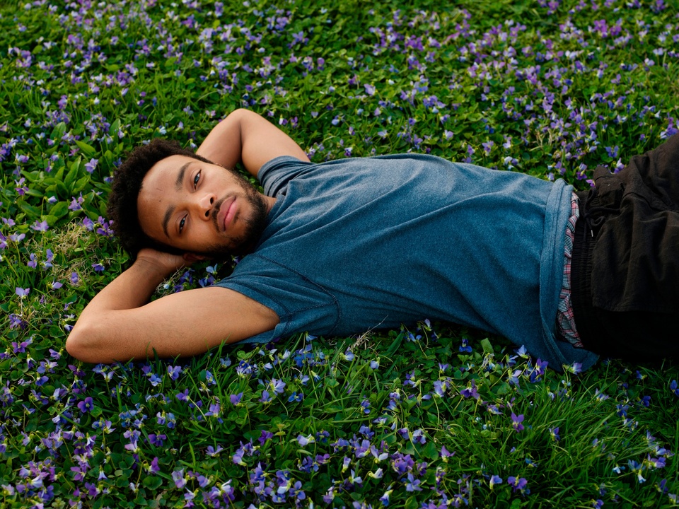 Inkjet print of a person lying in a field of green plants with small purple flowers, their arms behind their head, staring at the camera.