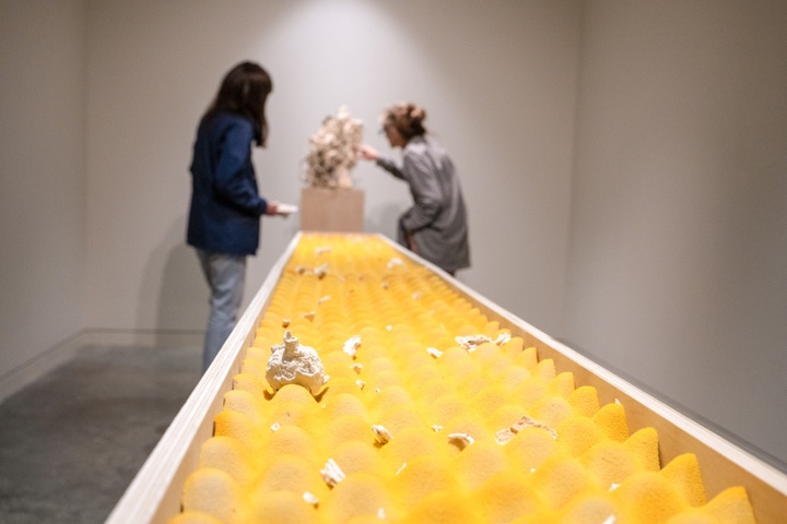 Low angle view of a long table-top sculpture. A strip of yellow egg-crate mattress topper rests in a trough. Nestled in the divots are small, cracked pieces of plaster. Two people look at a different sculpture in the background.