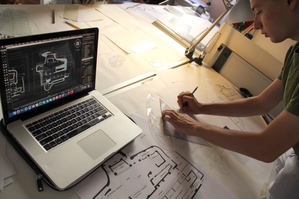 Person working on an architectural drawing referencing a computer on their work table.