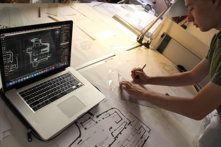 Person working on an architectural drawing referencing a computer on their work table.