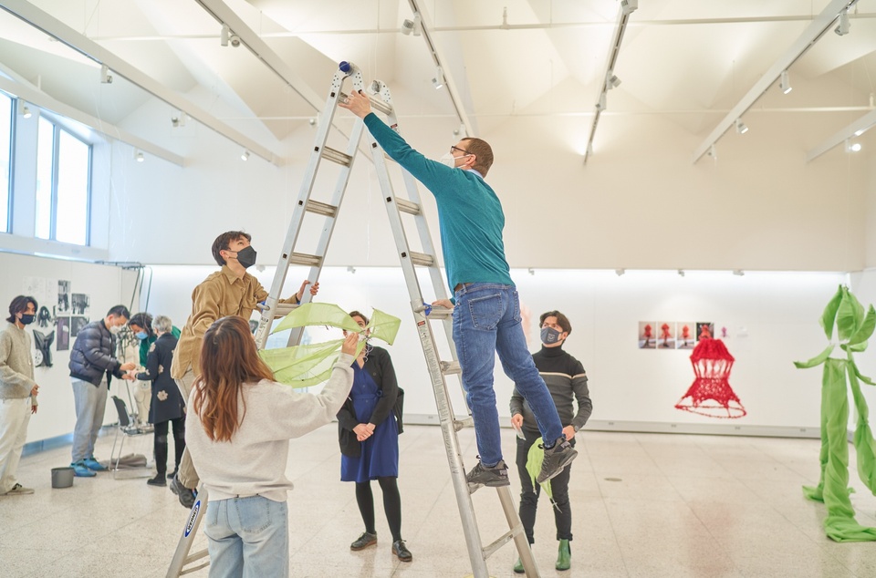 Two people climb a ladder to reach the ceiling of a gallery space. Colorful fabric constructions are hung around the room.