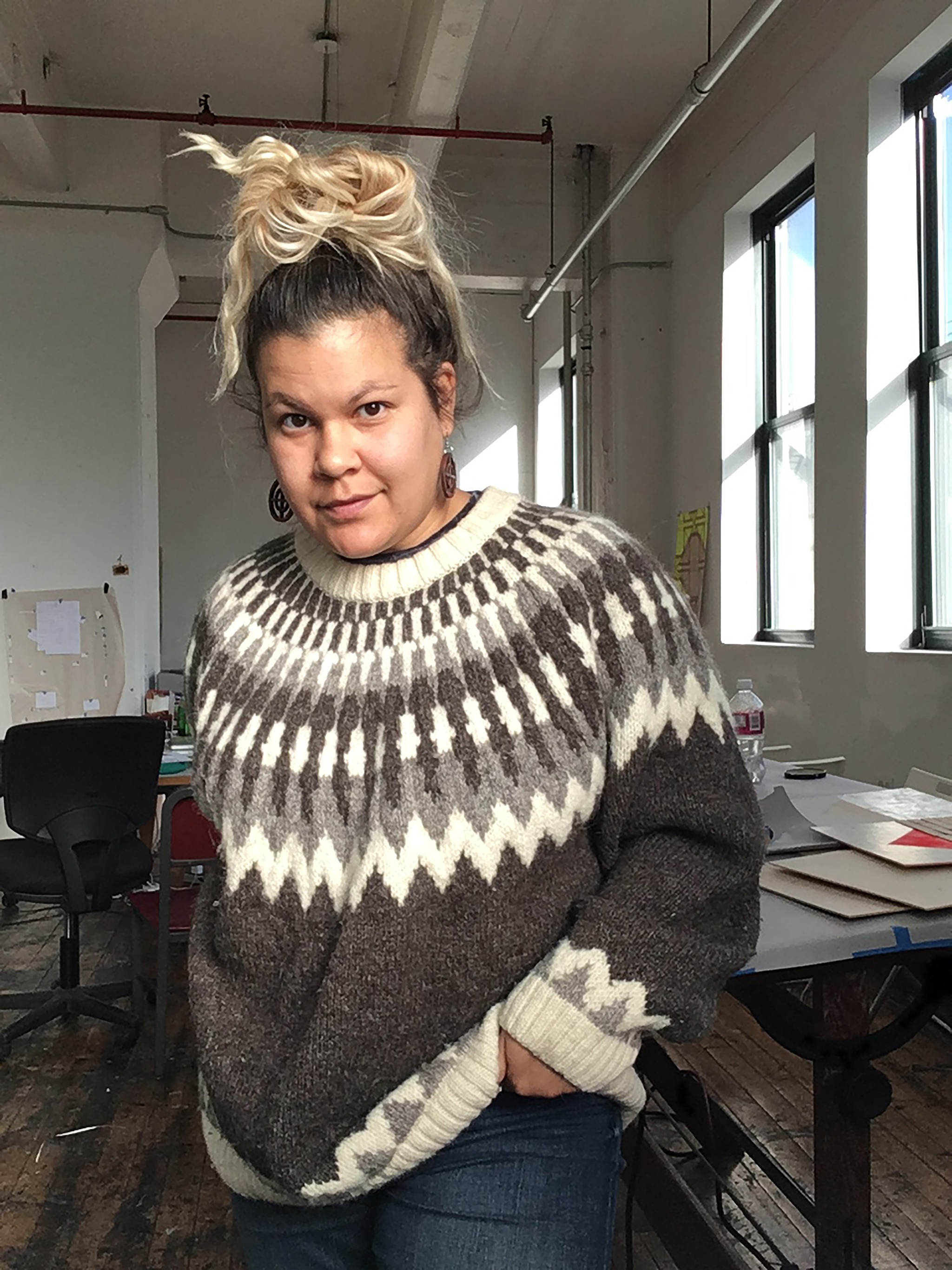 A photo of artist Heather Hunt standing with one hand in her pocket in what appears to be a studio or workspace. She wears an oversized sweater and has her two-tone dyed hair pulled up messily at the top of her head. 