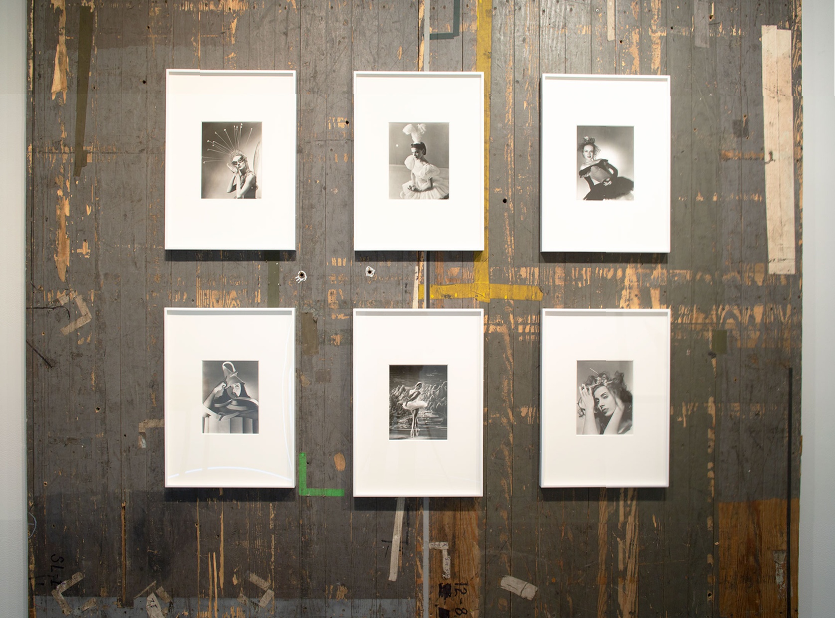 Six black and white photographs framed in white hang on a beaten up grey-brown hardwood floor, which also is adorned with tape and markings, presumably for dancers to mark their spots.