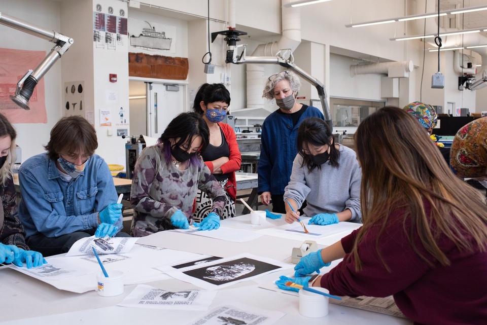 Students sit at a worktable in a printmaking studio, wearing blue gloves, handling sheets of artwork, while two instructors look on.