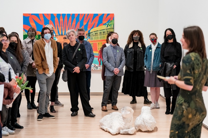 Person speaks to a crowd. On the floor between them is an indistinct, lifesize white sculpture of two sheep.