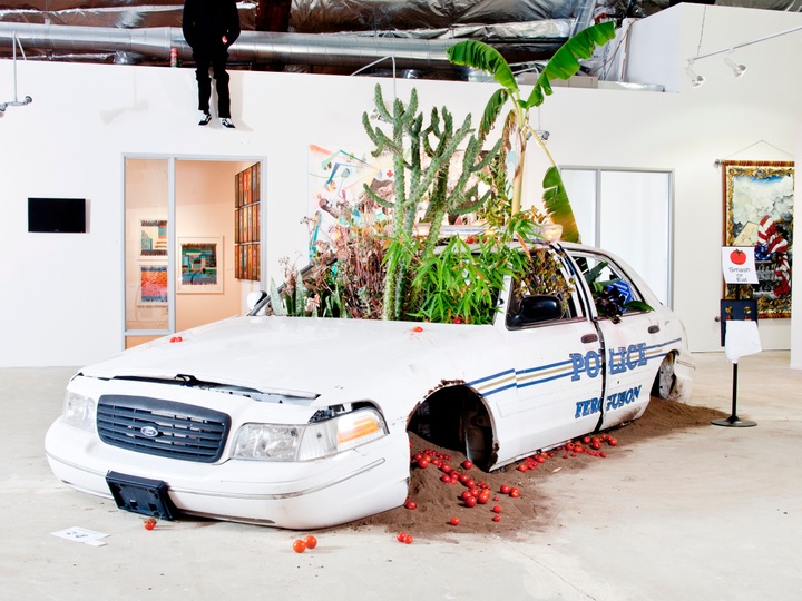 Installation of a white police car with blue detailing inside a white-box space. The car has no wheels, but tomatoes are strewn on the ground under the wheel wells, and green plants are growing up tall through the windshield.