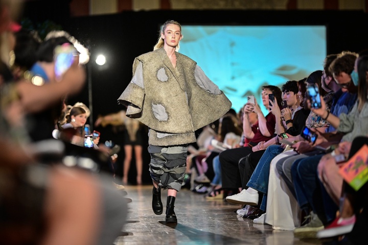 Model walks down a fashion show runway wearing a distressed burlap poncho and a narrow ruffled skirt of various denims.