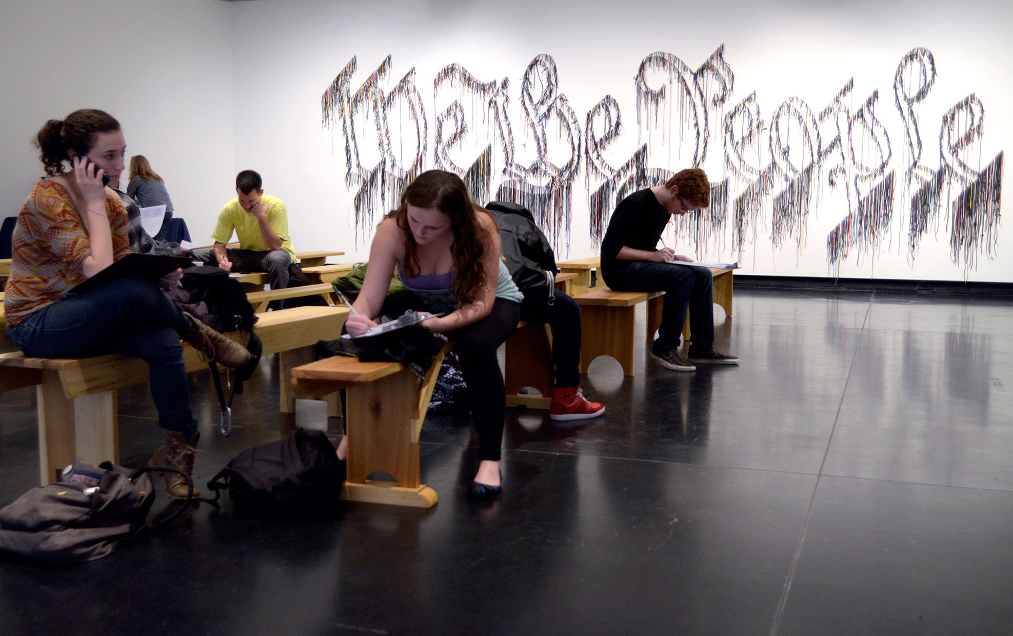 A group of young people sit on wooden benches. On the wall behind them, "We the People" is written with silver tinsel.