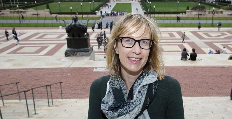 A white woman with dirty blond hair smiles with Columbia University's Morningside campus in the background. She wears glasses and her bangs are swept across her forehead. 