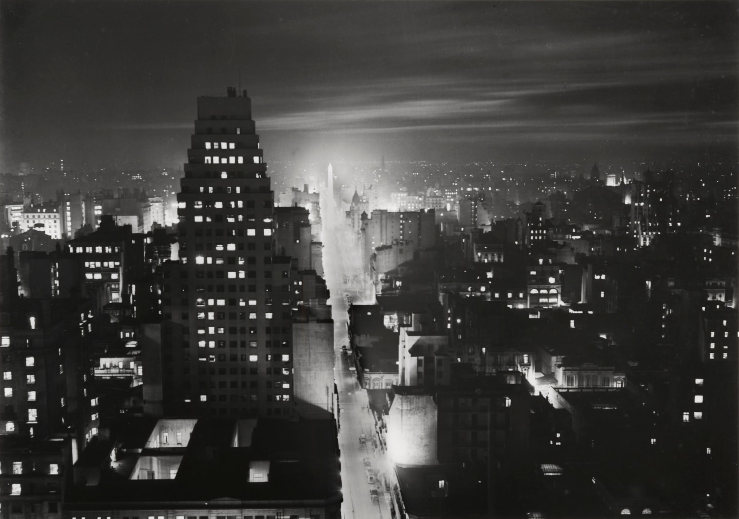 Black-and-white nighttime photograph of Buenos Aires's Corrientes Street, viewed from an elevated vantage point. Streams of headlights and illuminated storefronts line the broad avenue, while the city's buildings recede into the distance beneath a dark sky.