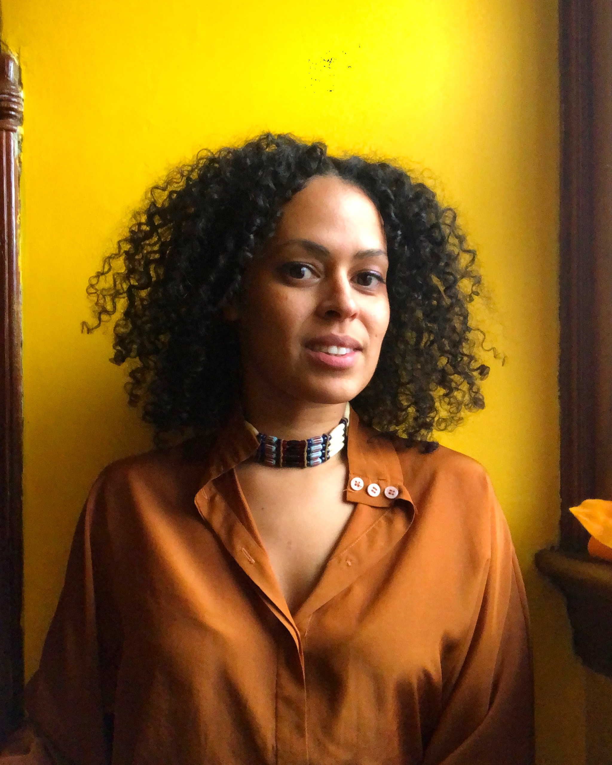 A photo of the artist Ana María Agüero Jahannes against a bright yellow background. Jahannes has curly hair and wears an orange-brown blouse and beaded choker. 