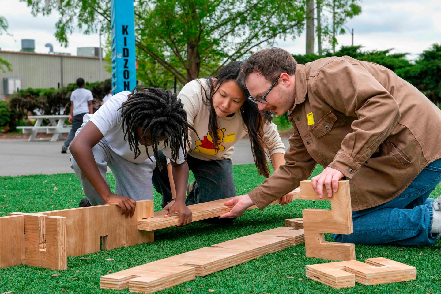 A man works with two students to build a modular wooden structure on a playground.