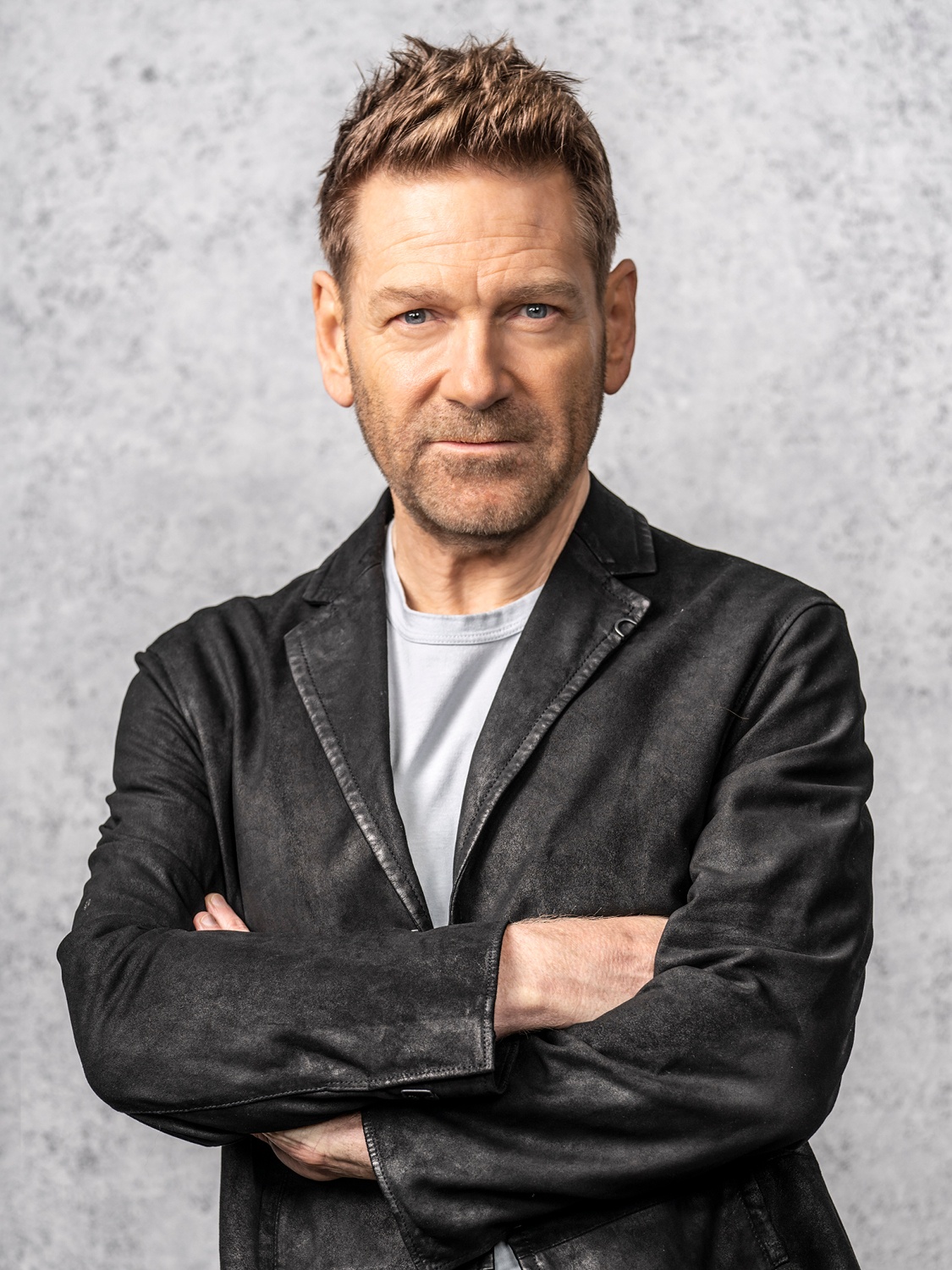 Kenneth Branagh, a white male with brown hair, stands center frame facing forward with his arms crossed. He stands in front of a blurred gray background, in an open black waxed leather blazer and light gray shirt.