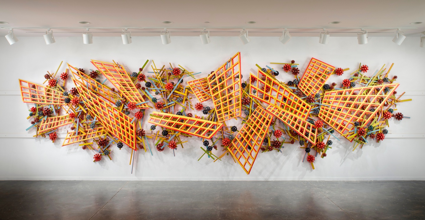 Wall hung abstract sculptural installation of skewed yellow/red lattice shapes with light blue and light green bars criss crossing through them. There are red and blue flower shapes throughout the sculpture.