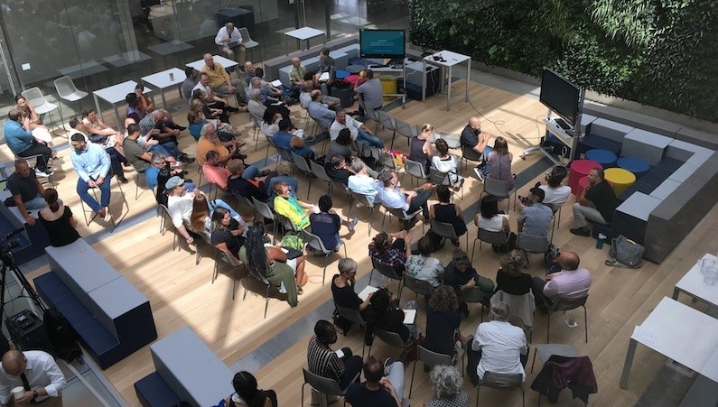 A large group of people photographed from above talk to each other in a sunny atrium. 