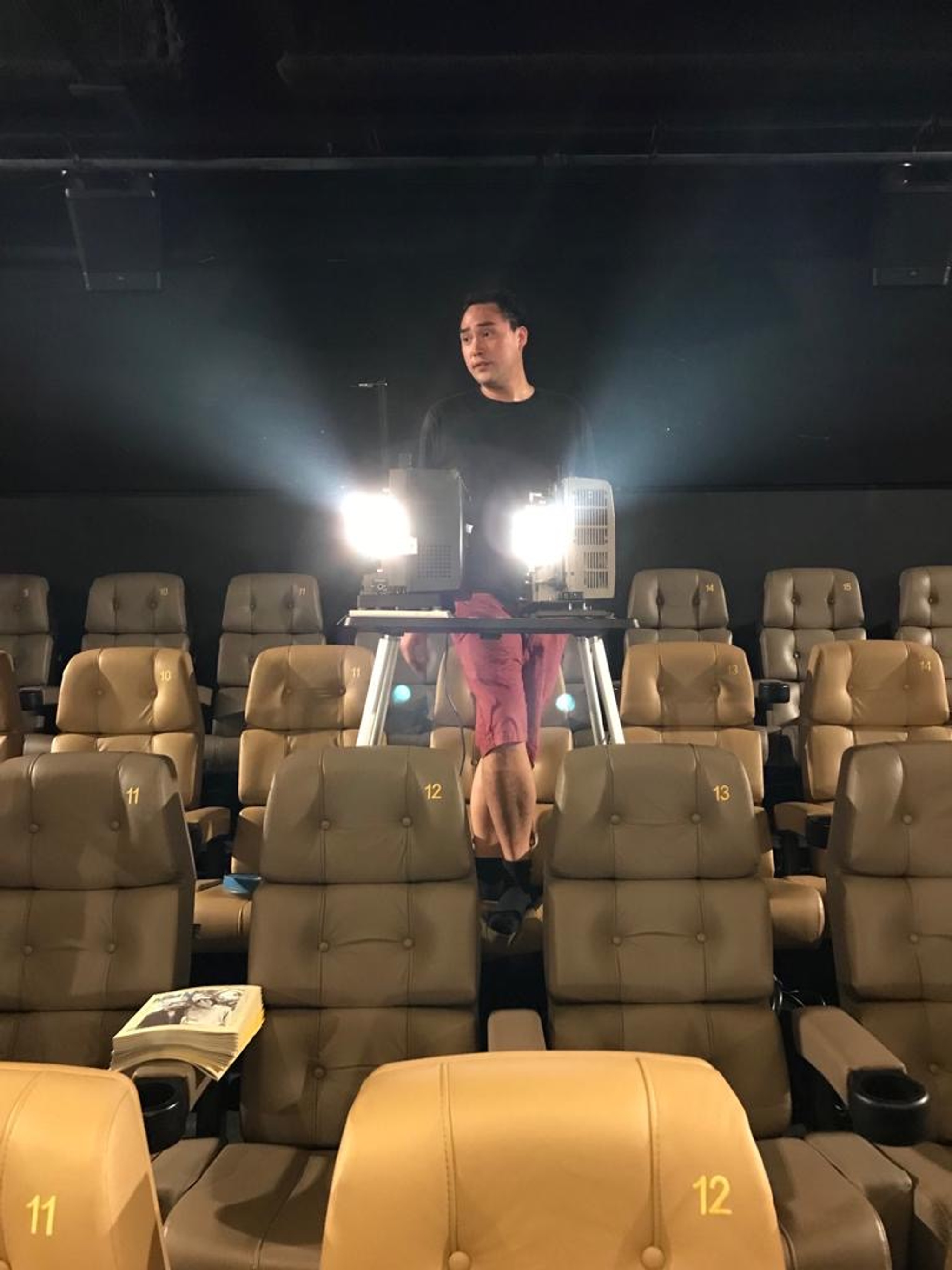  A photo of the artist Simon Liu in a black longsleeve shirt and red shorts, standing behind a projector and rows of seats in a movie theater.