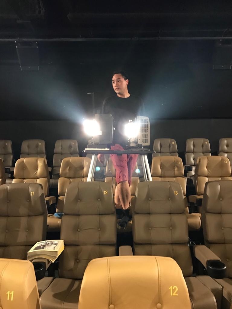  A photo of the artist Simon Liu in a black longsleeve shirt and red shorts, standing behind a projector and rows of seats in a movie theater.