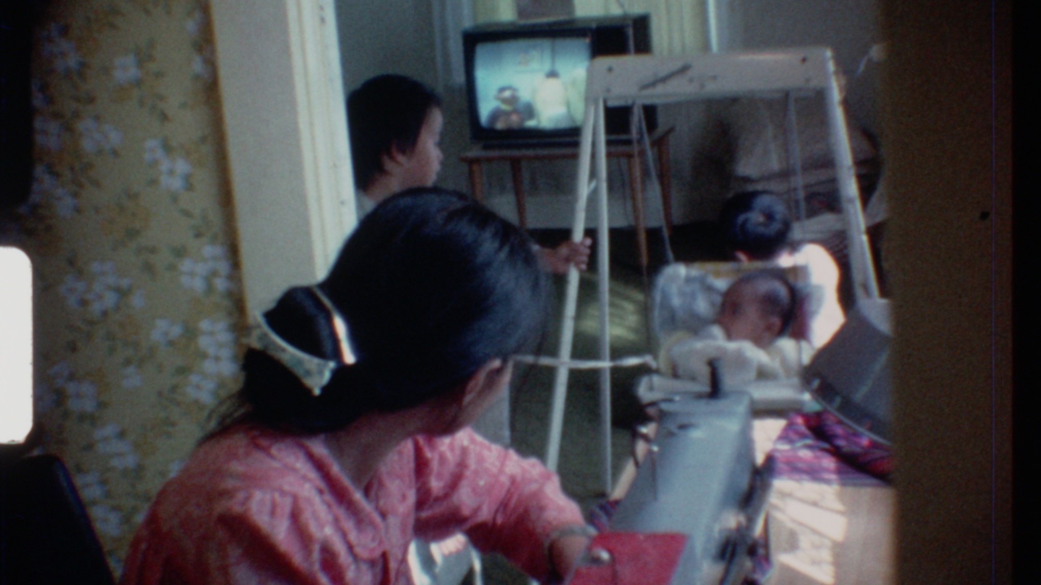 A Vietnamese woman sits at her sewing machine, facing away from the camera, her smooth black hair gathered in a low bun, fastened with a gold clip and white, bow-shaped barrette. The woman’s face is turned toward the living room toward three children: a baby in a swing, pulled by a rope in the hand of a young boy in profile, and the back of a young boy’s head. On a 13 inch box television,  which is perched on a wooden table, Burt and Ernie from Sesame Street are on the screen.