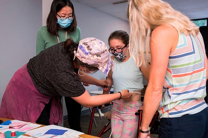 A student with a tape measure measures the waist of her model with the help of another student and the model's mom.