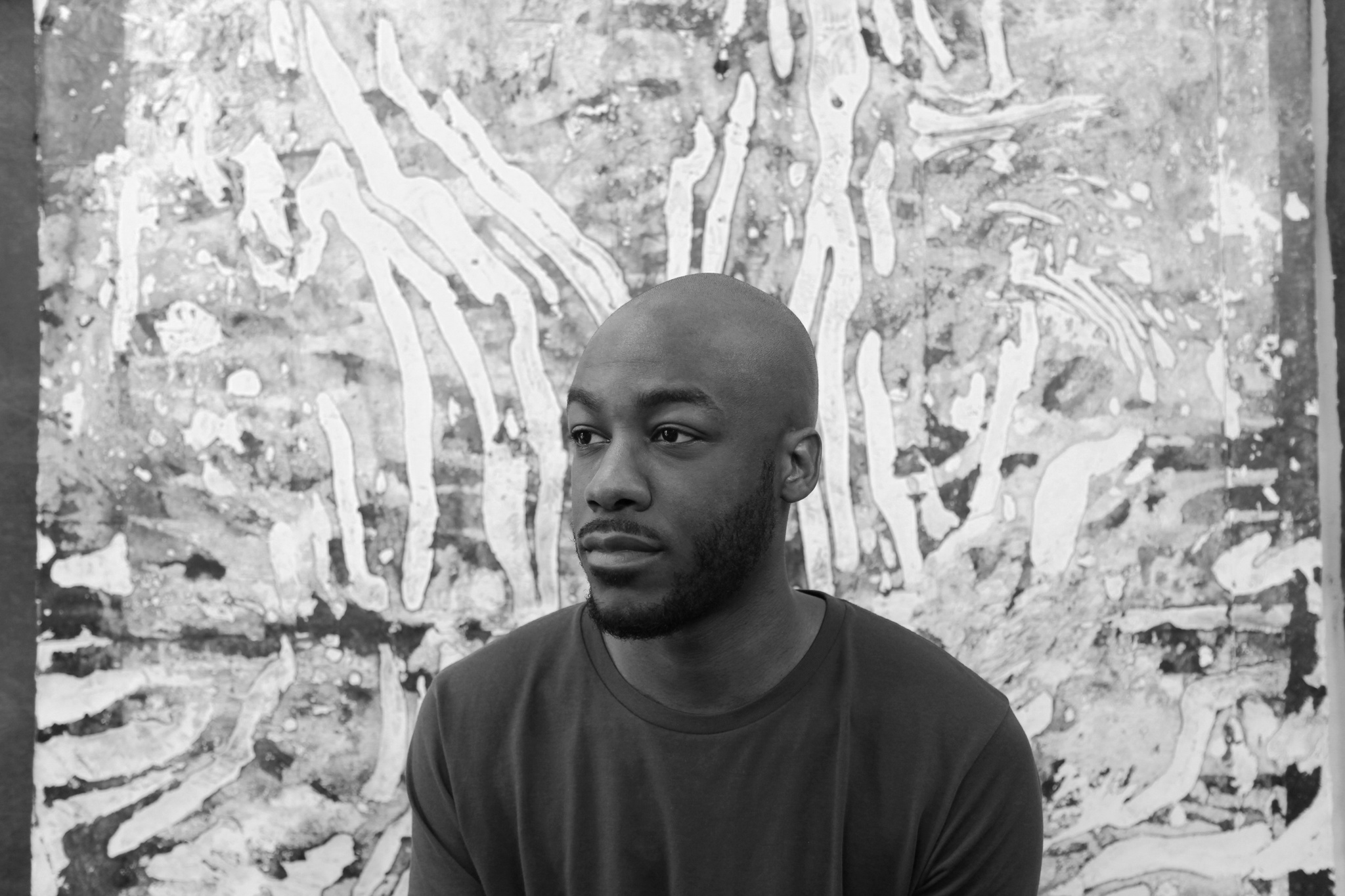 A photo of the artist Tajh Rust sitting in front of an abstract painting. Rust has a beard and looks away from the camera out of the frame of the image.