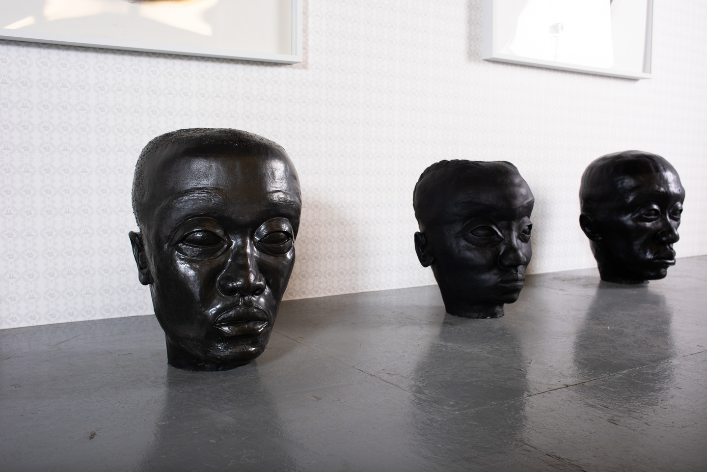 Three black, shiny sculptural heads with well-articulated eye sockets sitting in a row on the floor. 