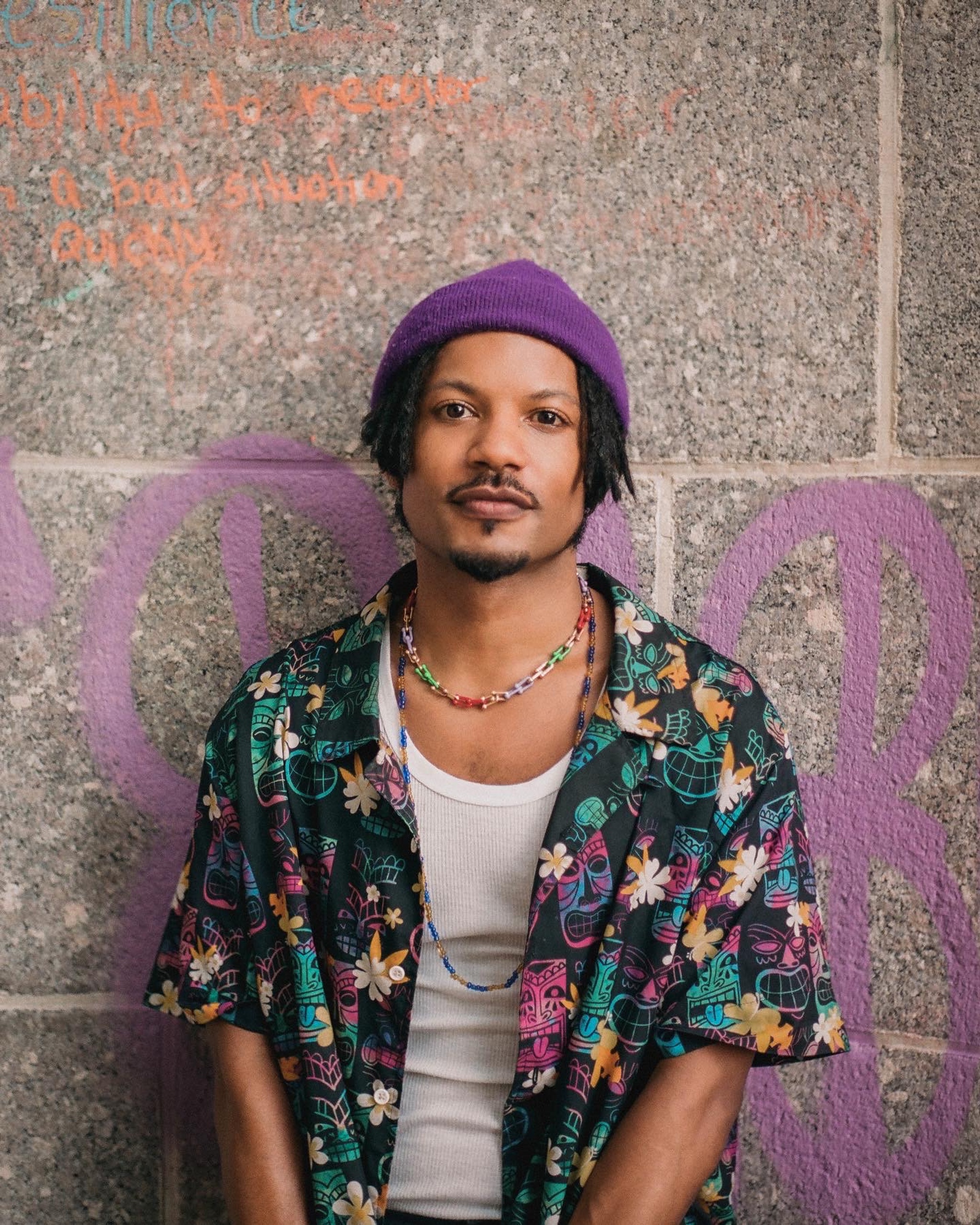 Artist Jaime Cepero looks directly at us in a portrait posed against a stone wall. An Afrolatino person, they wear a purple cap over short locs and a colorful floral print shirt. They have a contented look on their face. 