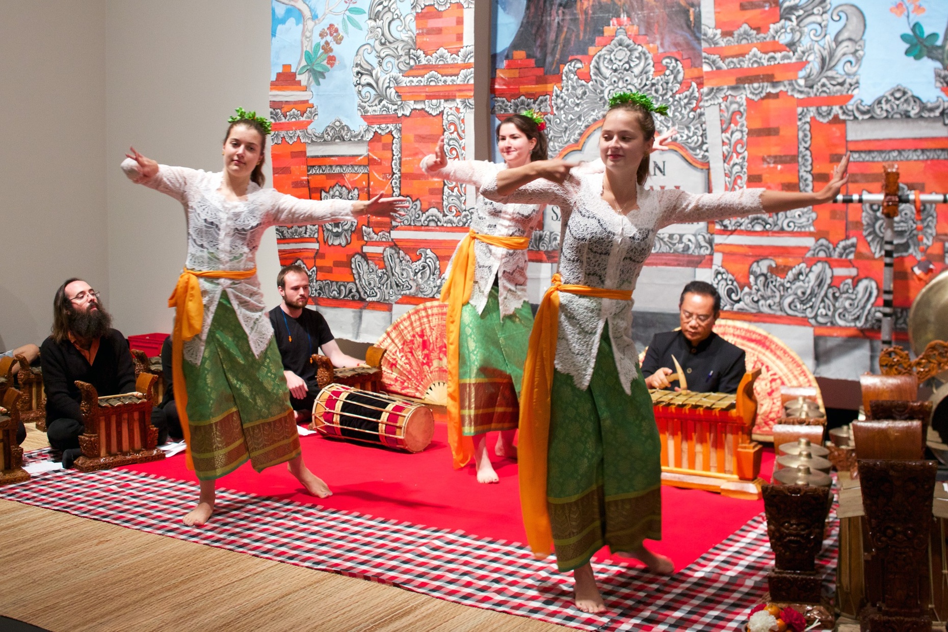 Three, light-skinned, female dancers in oriental-style clothing posing on a red rug with different small instruments lined up behind and around them. A tanned, Asian man sits playing a xylophone type instrument behind the dancers in front  of elaborate wall hangings depicting a red temple.