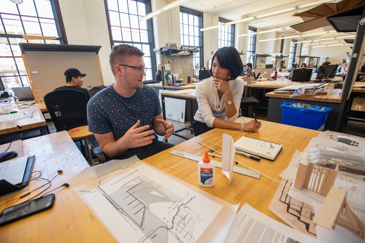 Professor consults with a student at their L-shaped desk in a large studio space filled with other private desks.