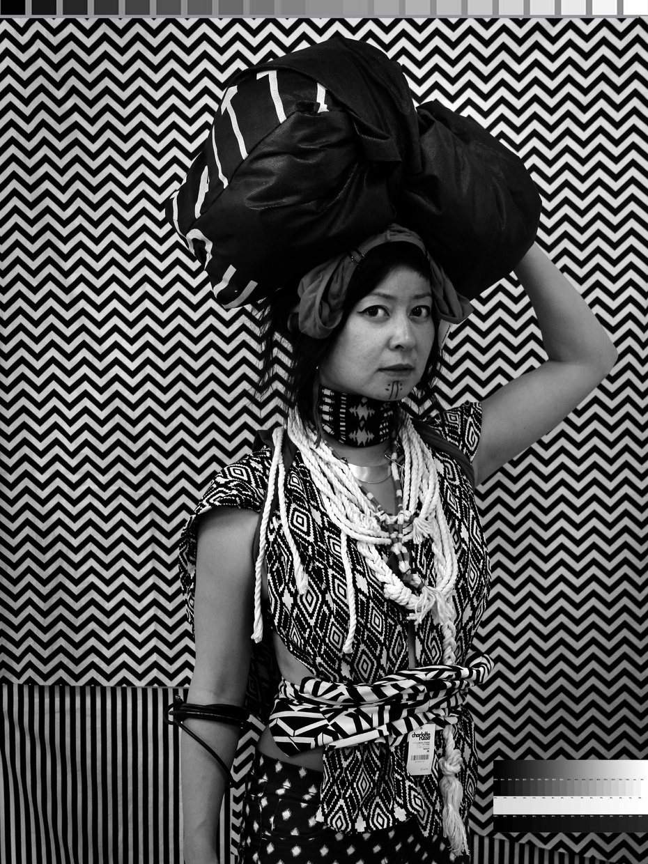 A black and white photograph of a young, Asian women holding a black bundle on her head wearing traditional dress in front of an abstract background.