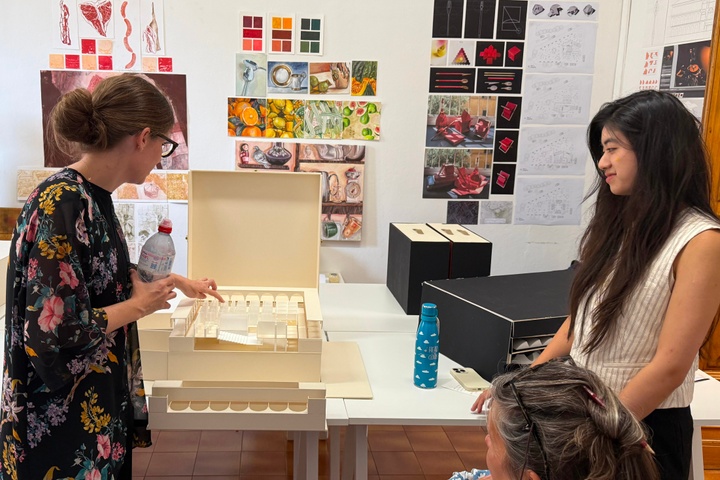 Students gather around a table with white architectural models. one student is presenting their project.