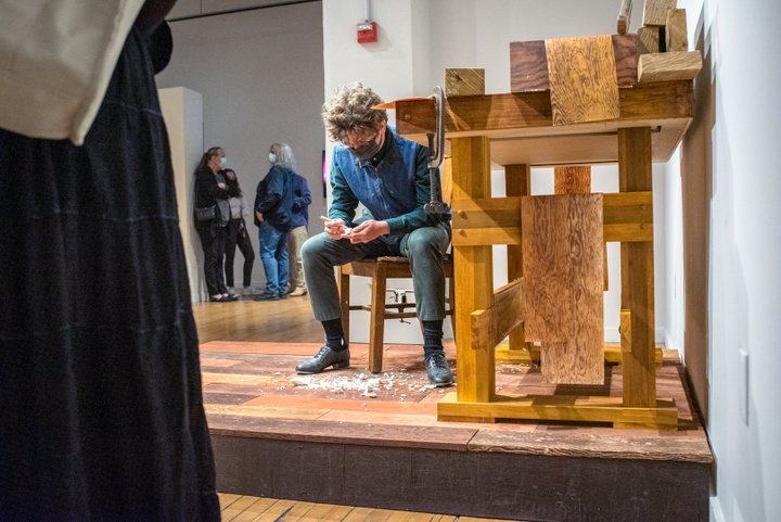 Person sits in a wooden chair at a carpenter's bench on a short wooden dais. They lean over their knees and hand-carve a small piece of soap. People stand around the dais and watch.