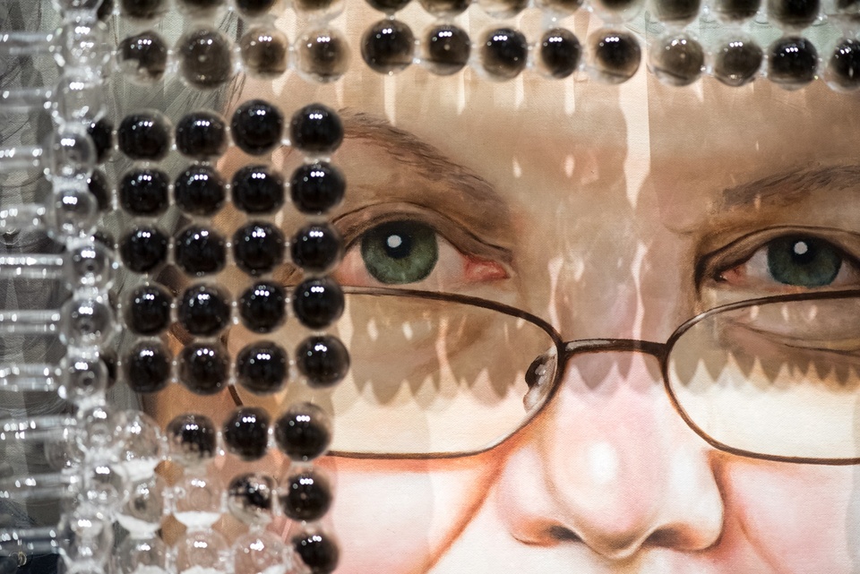 Detail of painting of an older light-skinned woman's face. Shading her eyes are rows of small glass pipes with char on the bottom.