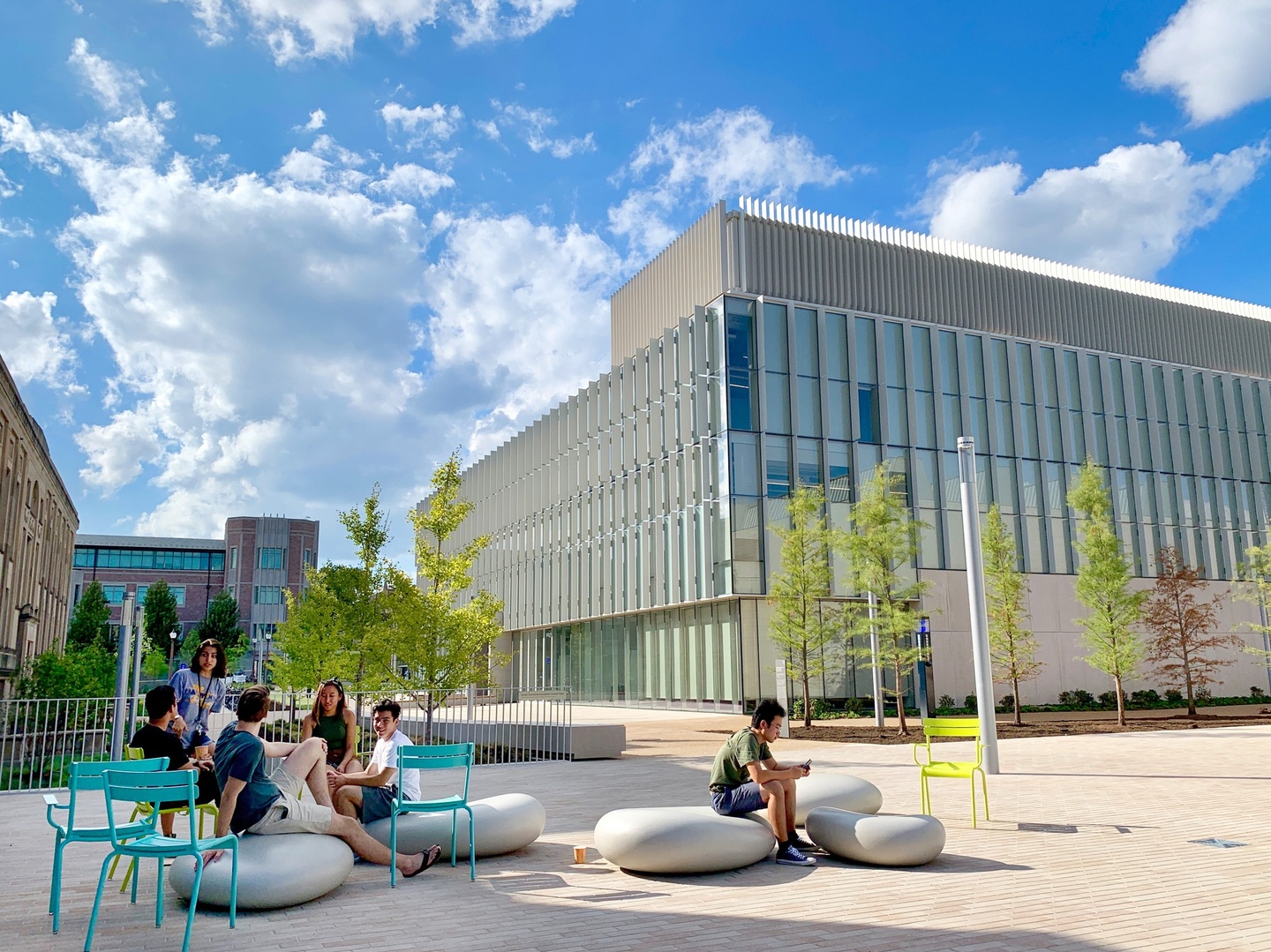 Bricked patio in front of a modern glass walled building. People sit on large smooth concrete boulders and turquoise metal chairs.