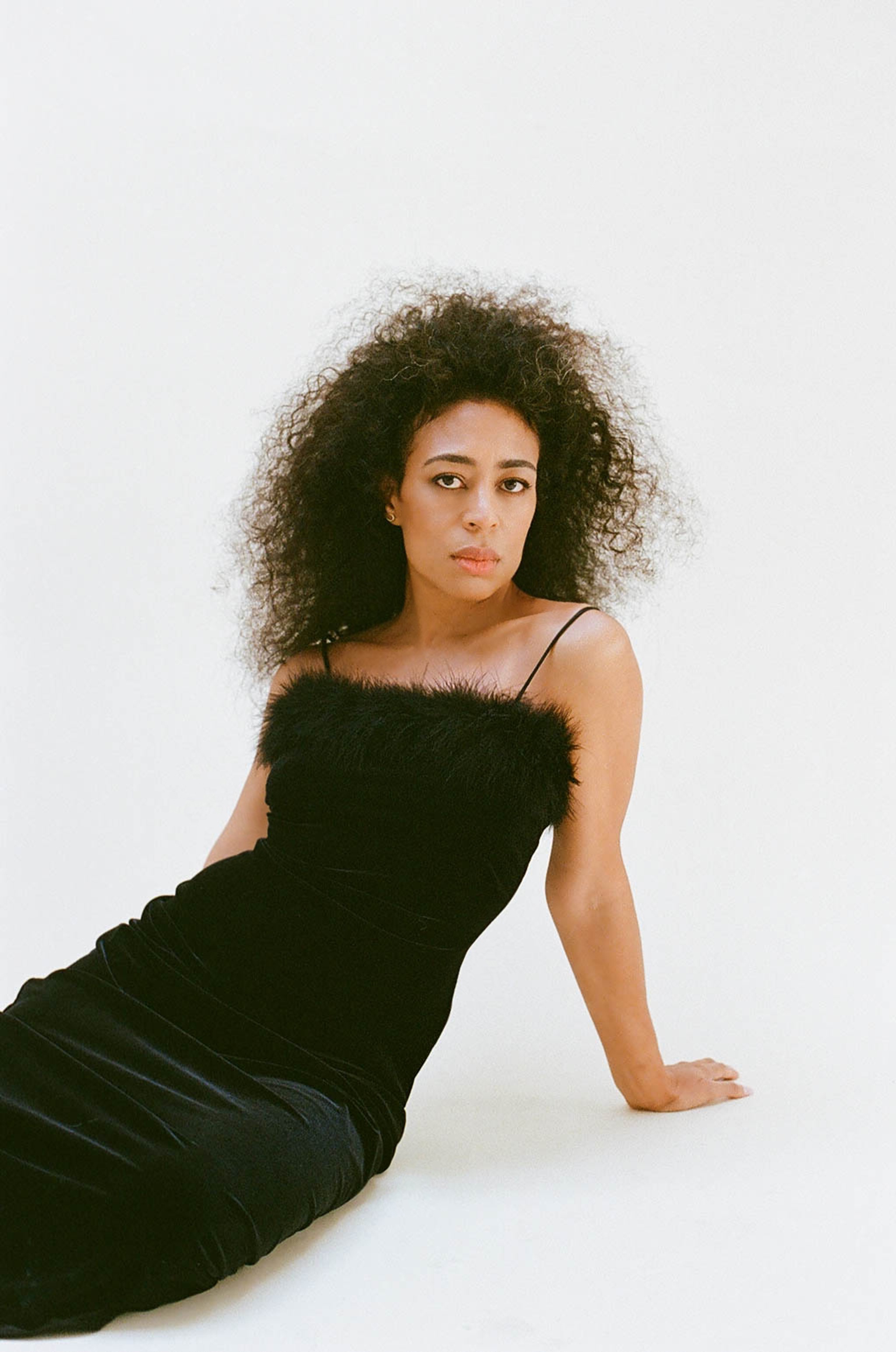 The artist Candice Hoyes, a Black woman with light brown skin and curly brown hair, reclines against a white backdrop. She wears a long black, velvety dress with a band of black fur across the chest. She looks directly at us with a focused, neutral expression.