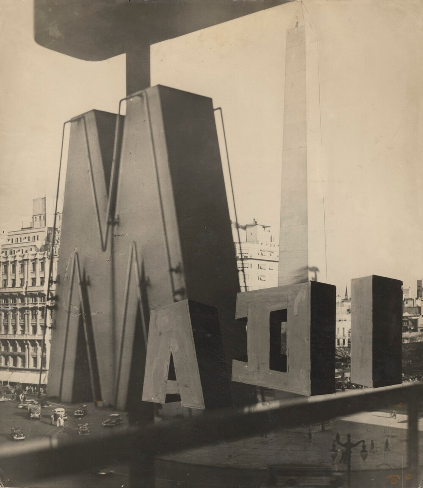 A black-and-white photograph showing large geometric letters and abstract sculptural forms appear to be on a rooftop, with a tall obelisk and city buildings visible in the background. Cars and pedestrians can be seen on the street below.