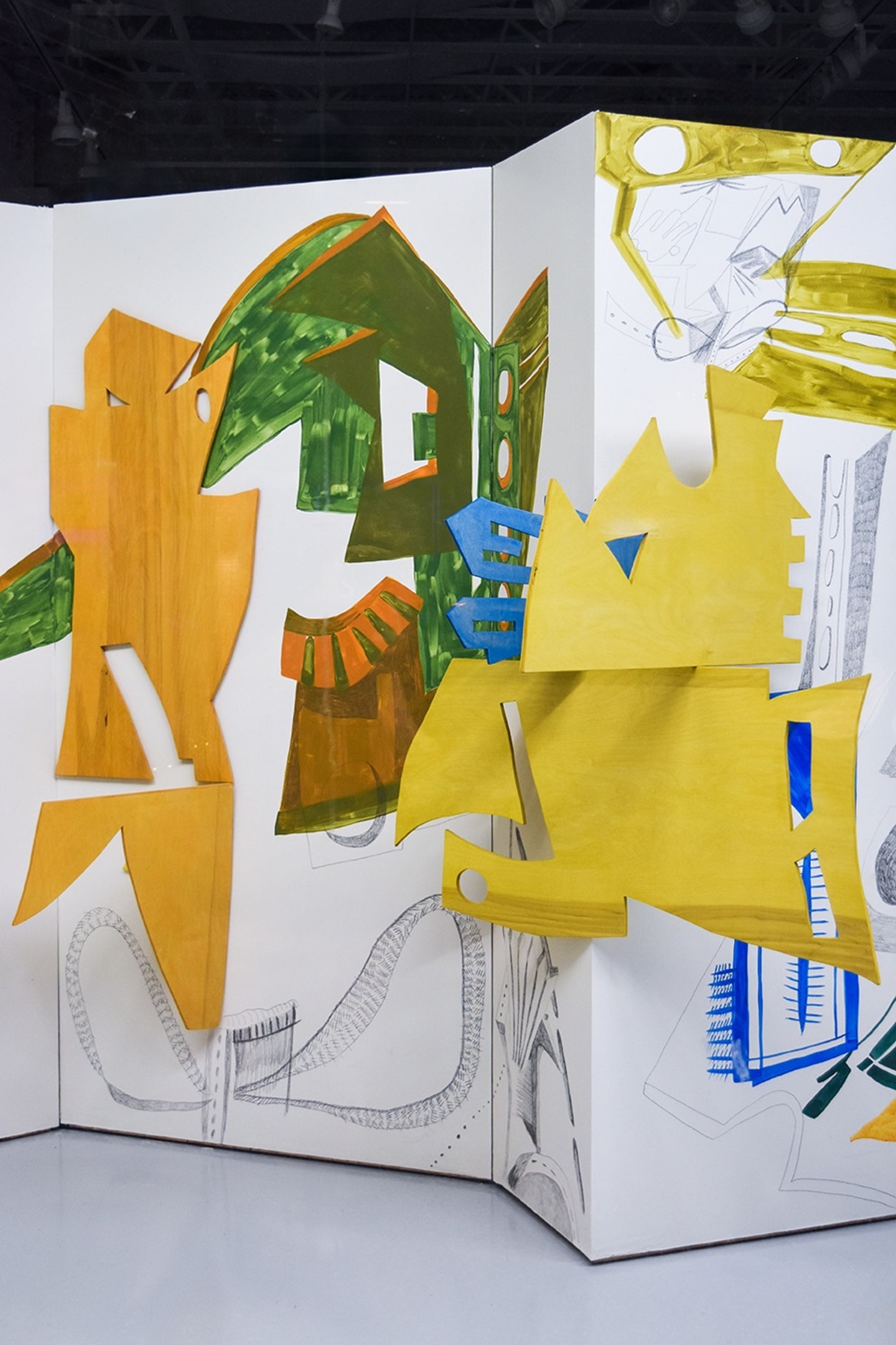 An abstract, mixed-media installation by Ribaudo featuring layered yellow, green, and blue painted wood cutouts mounted on multi-paneled white gallery walls with graphite line drawings