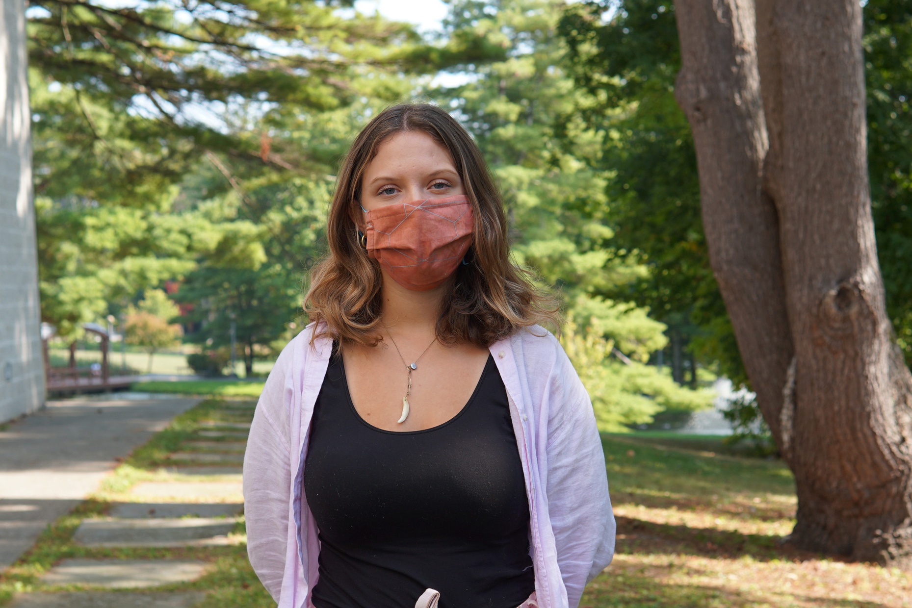 A light-skinned, young female wears a red mask with gray patterns printed on it standing in front of trees.