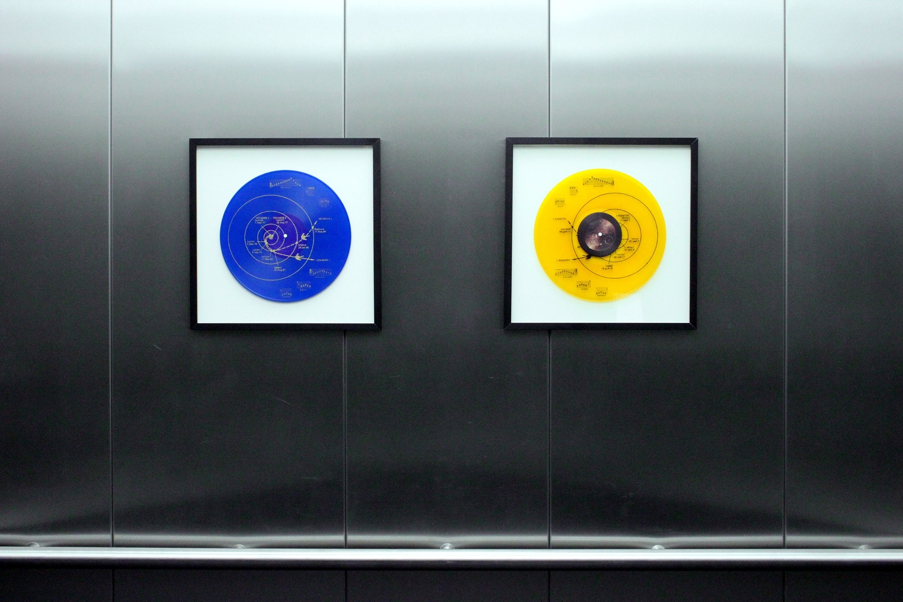 Two vinyl records, one blue and one yellow, hang in black frames on a metal wall.
