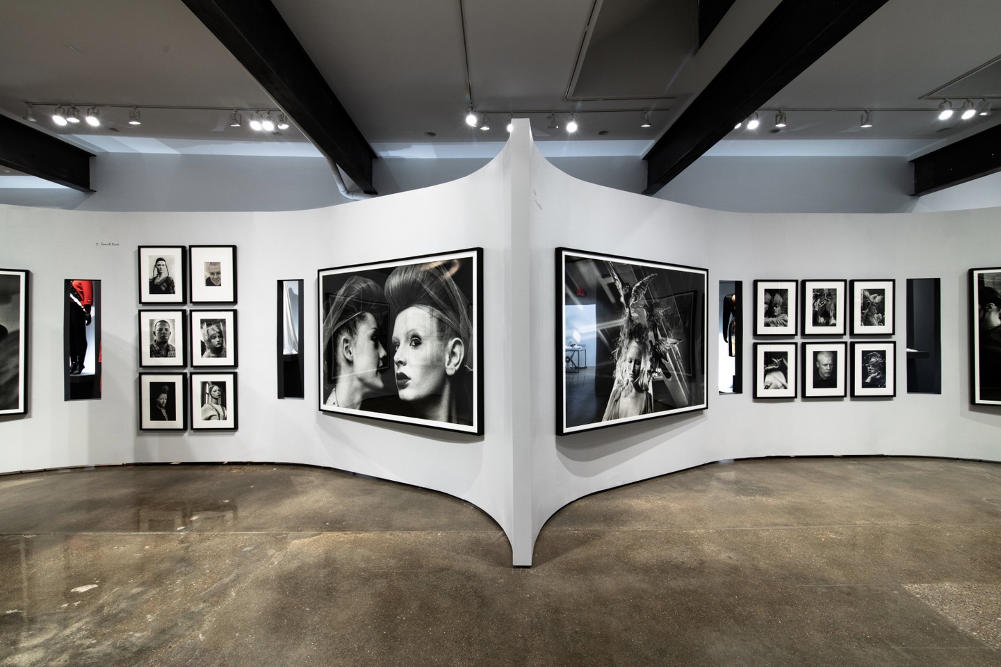 Interior installation view of an artwork exhibition, hung on a pair of curved walls that meet in the middle. Most of the framed works are black-and-white photographs.