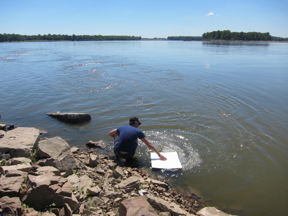 A figure sitting by a bank pressing a large white rectangular sheet over a body of water