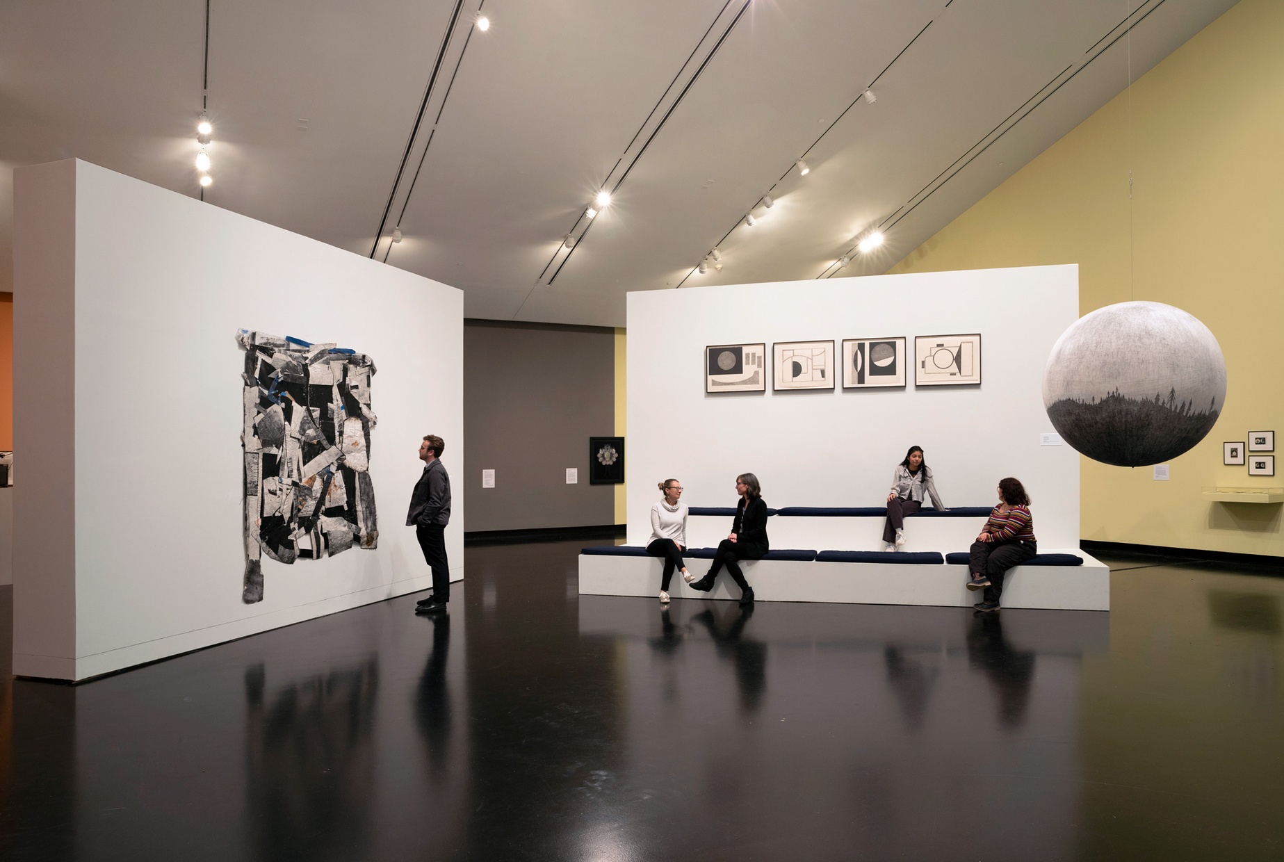In a gallery, a man stands to our left observing an artwork on a freestanding wall, while four people to our right sit on benches in front of a freestanding wall.