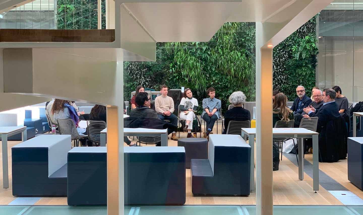 A large group of students and faculty sit in a circle of chairs in an atrium-like room. Behind the group is a two-story, living green wall.