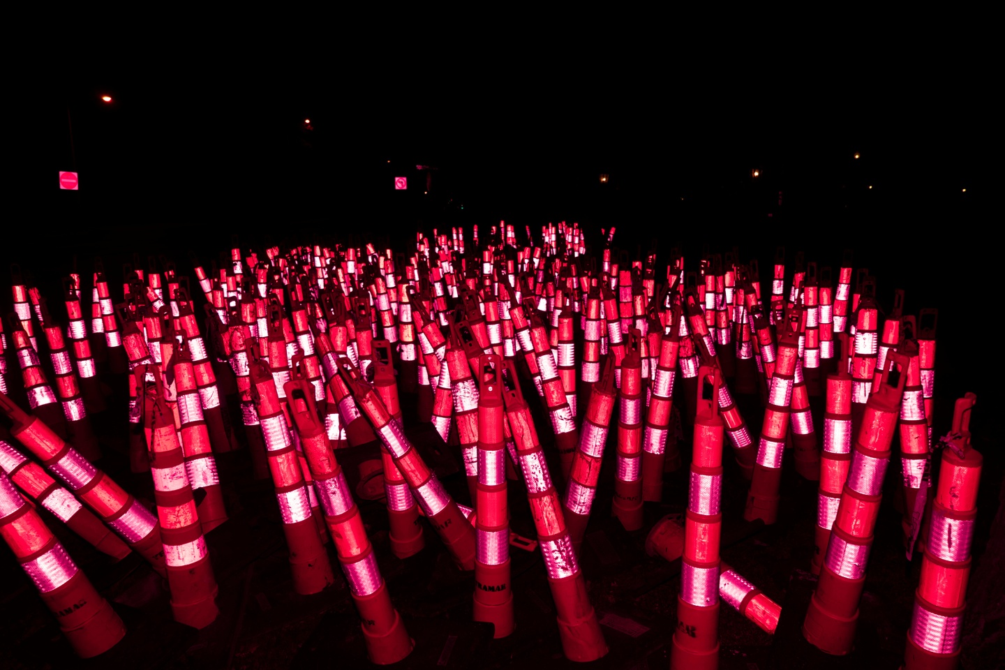 Nighttime photograph of a cluster of hundreds of orange safety cones photographed with a red light, causing the reflective stripes to glow eerily.