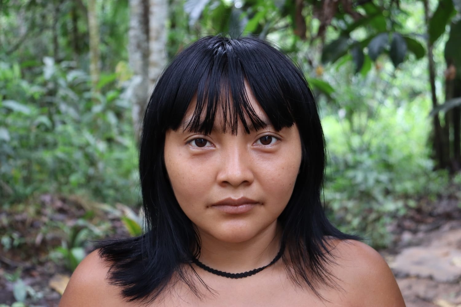 A portrait of artist Aida Harika. An Indigenous Yanomami woman with shoulder-length straight dark hair, Aida wears a thin choker and stands in front of a background of lush green leaves and trees. 