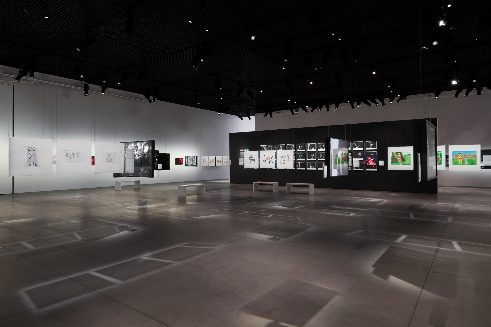 An art gallery with drawings and photos on walls and suspended from the ceiling. The suspended artworks make shadows of intersecting lines on the gray concrete floors. In the back of the gallery is a long wall painted black with a grid of black-and-white photo portraits of Yanomami subjects.