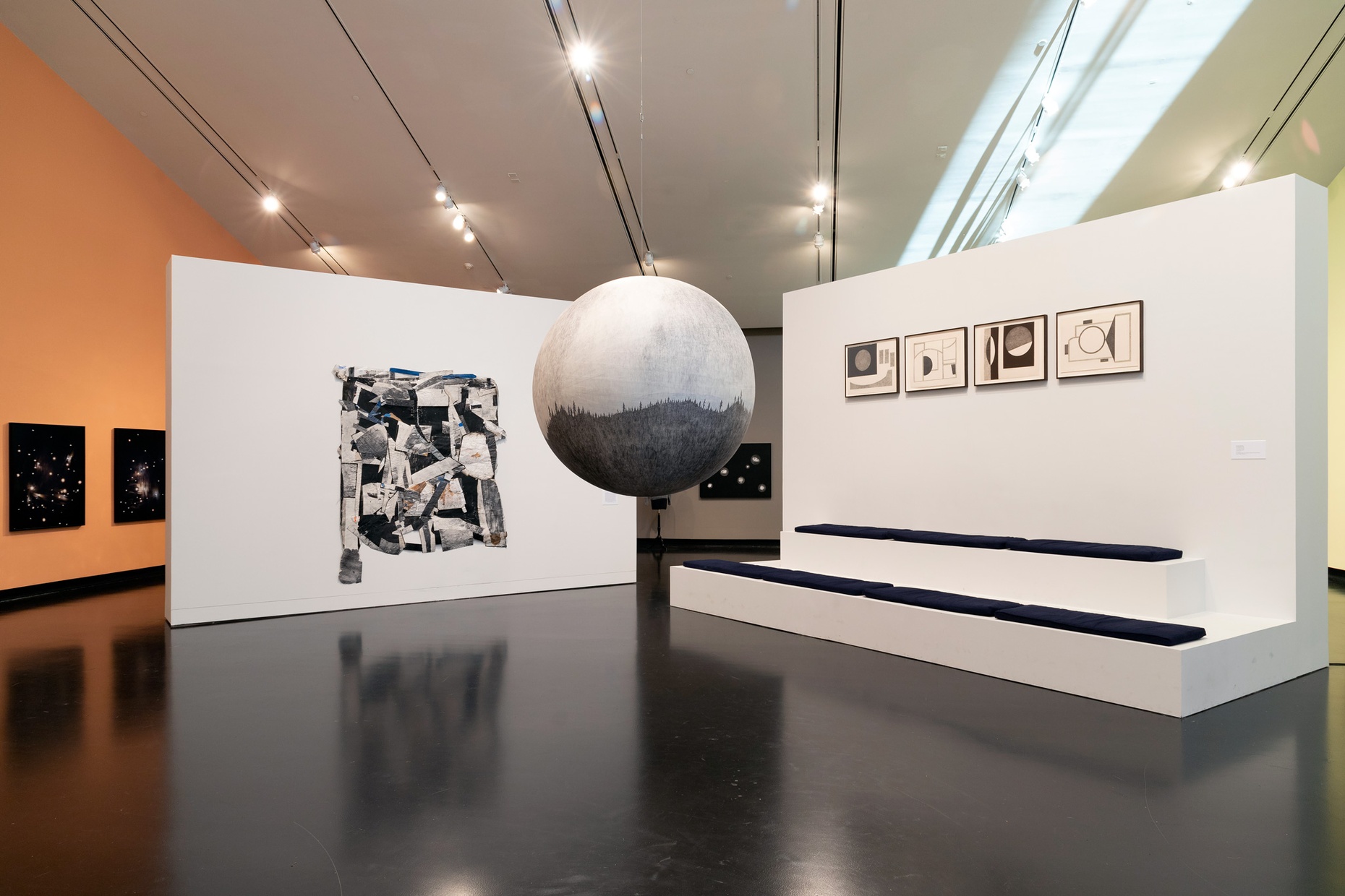 In a gallery, two freestanding white walls, featuring hanging artworks, are angled towards the center of the image where a light and dark gray sphere hangs from the ceiling.