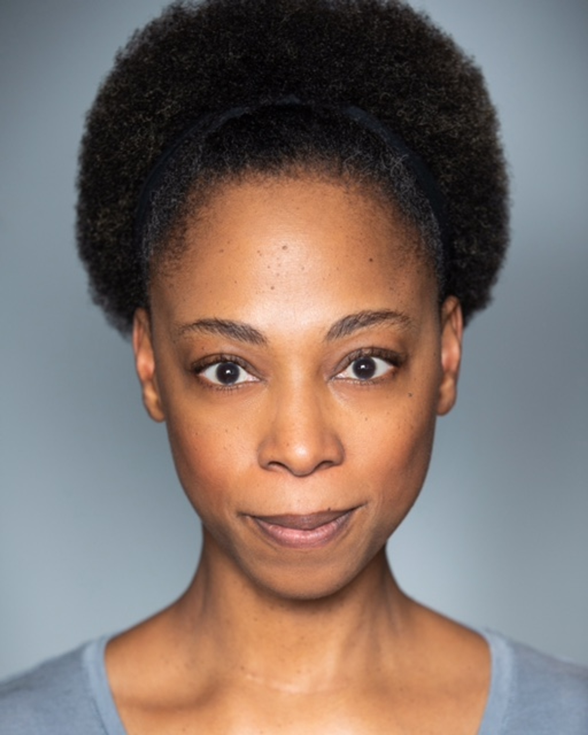 A portrait of actor Alana Maria, a Black woman with her hair pulled back so it fans out behind her head. She smiles slightly without opening her mouth.