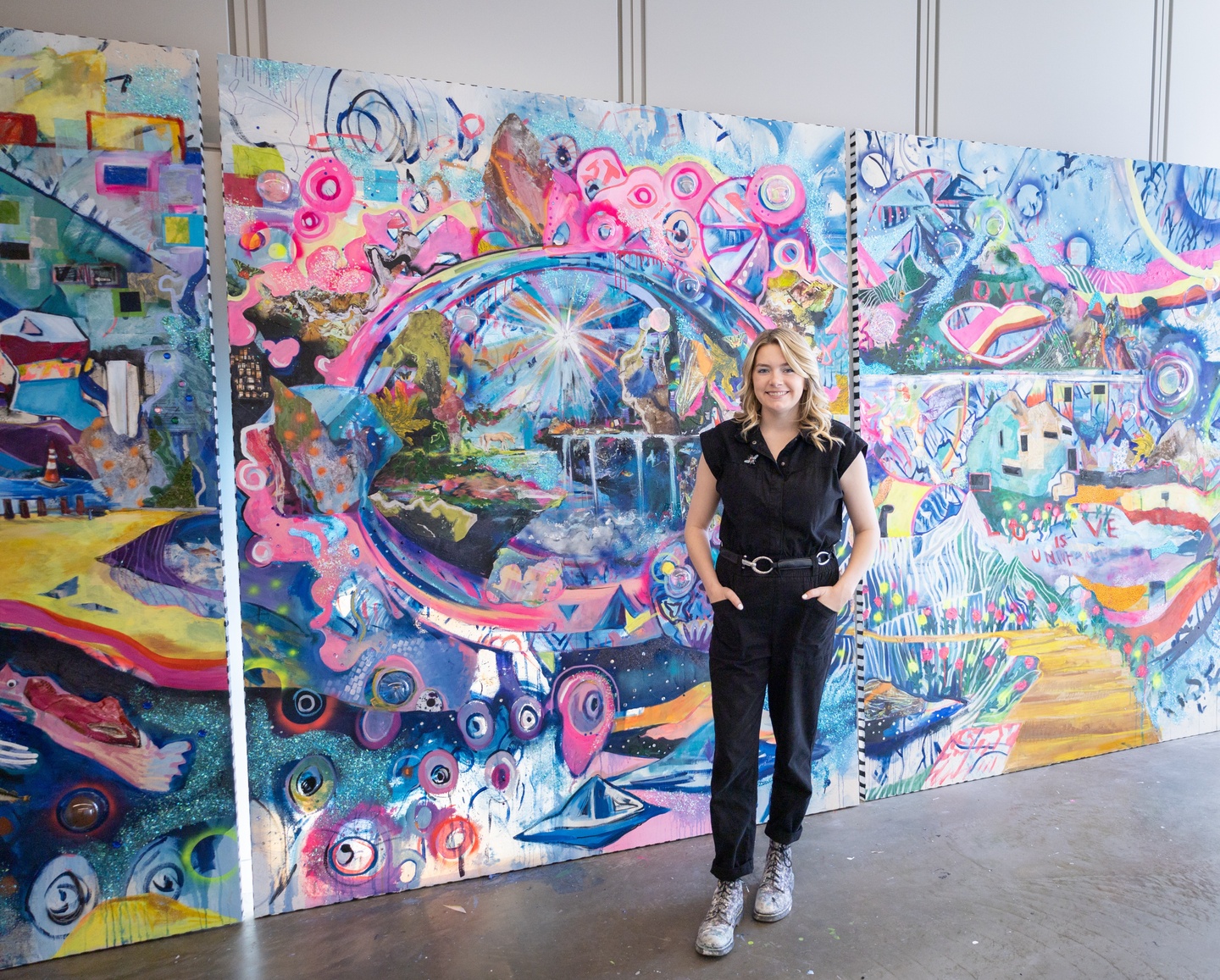 woman with long blonde hair wearing black standing in front of 3 paintings taller than her