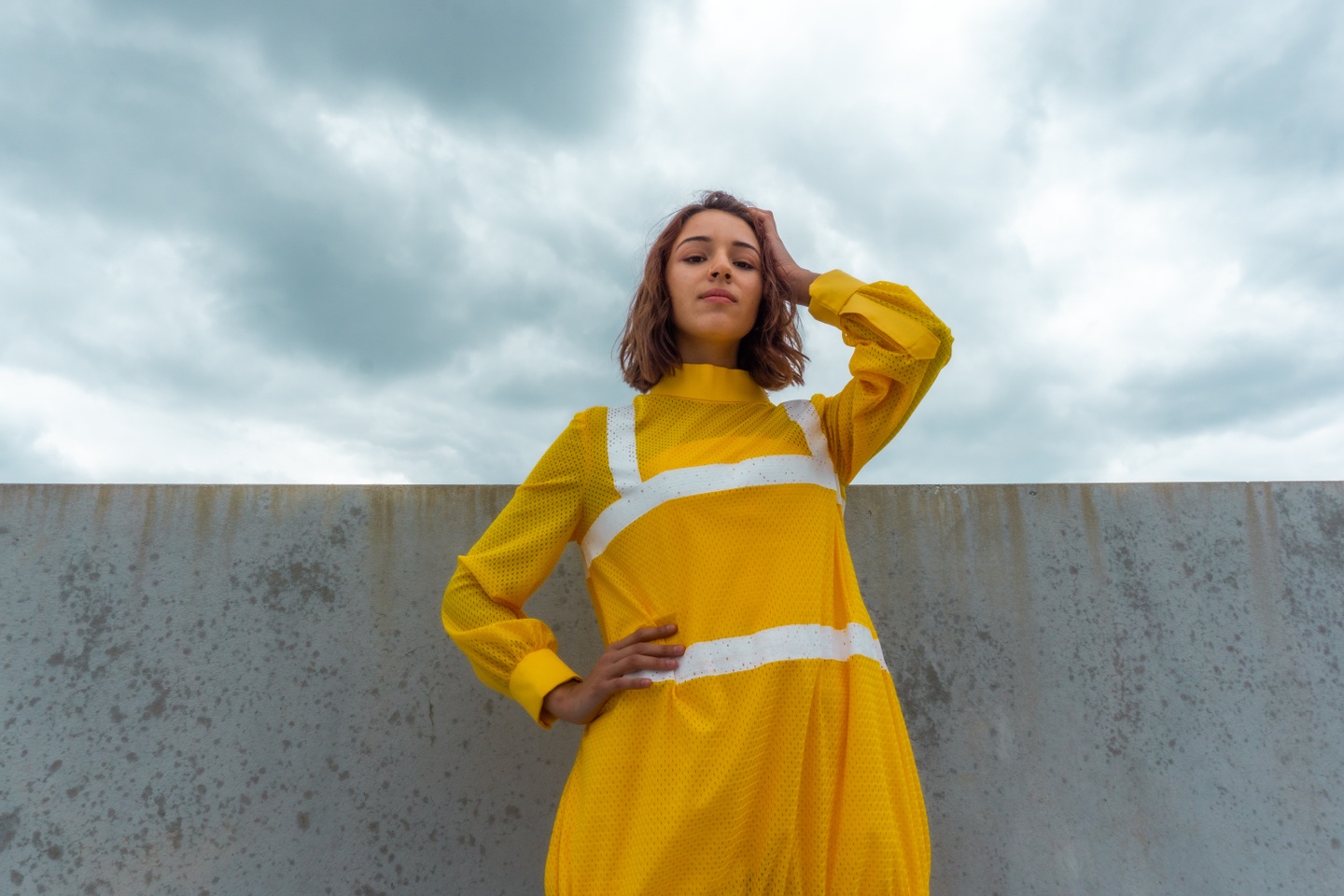 Model in a high-collared long-sleeve dress made of yellow mesh fabric similar to an athletic jersey. The dress has white paint striping across the shoulders, waist, and chest similar to a safety vest's reflective strips.